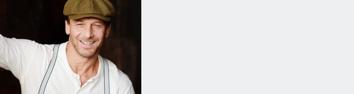 Schauspieler Hendrik Duryn mit weißem Hemd und brauner Mütze, lächelt in die Kamera.