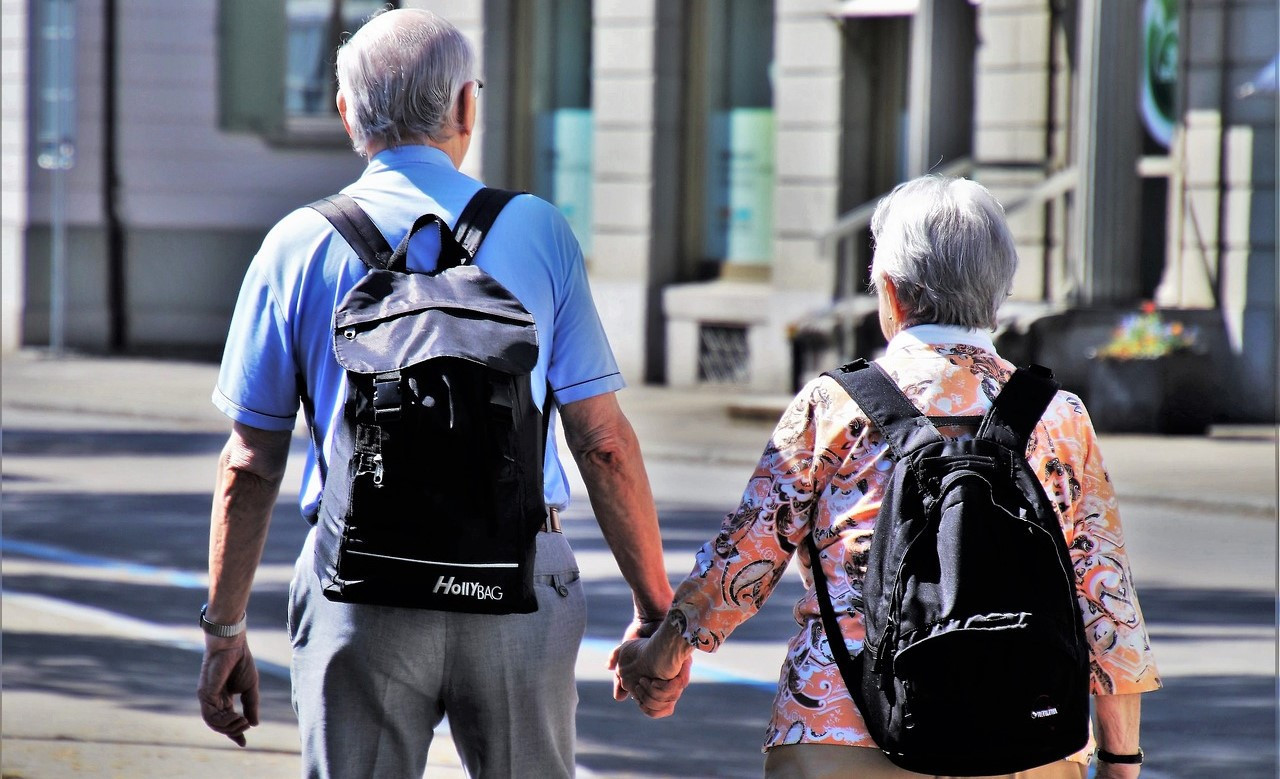 Ein älteres Paar Senioren mit schwarzen Rucksäcken, von hinten, Händchen haltend.